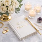 Agendă elegantă cu titlul „Tu Ești Magie” pe o masă decorată cu lumânări parfumate, cristale, trandafiri albi, bijuterii și un stilou auriu în formă de baghetă magică – simbol al feminității, rafinamentului și magiei interioare