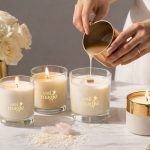 Femeie turnând ceară naturală în recipiente elegante pentru a crea lumânări handmade, alături de lumânări aprinse, cristale și flori albe – simbol al magiei lumânărilor făcute manual.